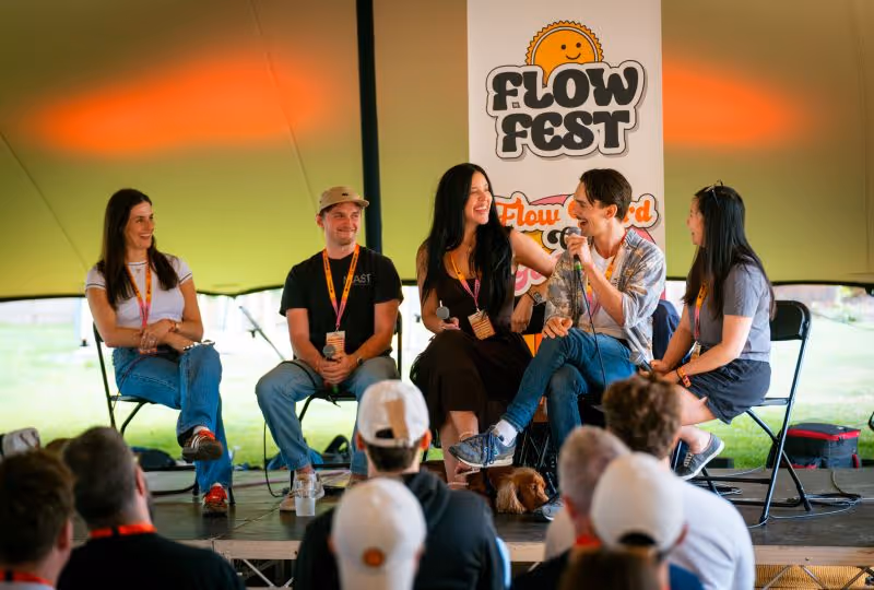 Five people sitting on chairs on a stage with microphones, talking and smiling at an event called Flow Fest, with an audience watching.